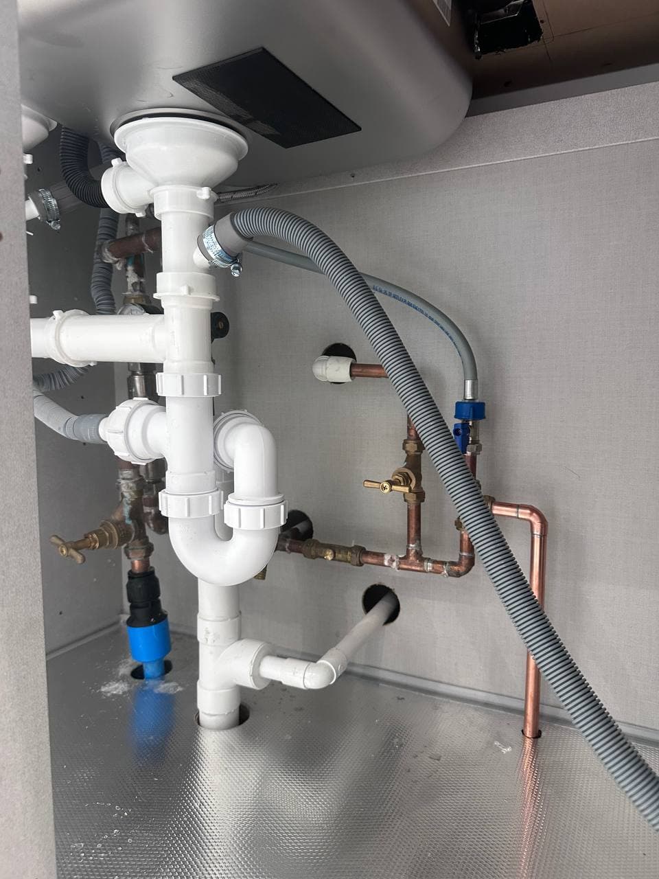 Plumbing under a kitchen sink: white waste pipes, copper pipework, isolation valves and a washing machine hose