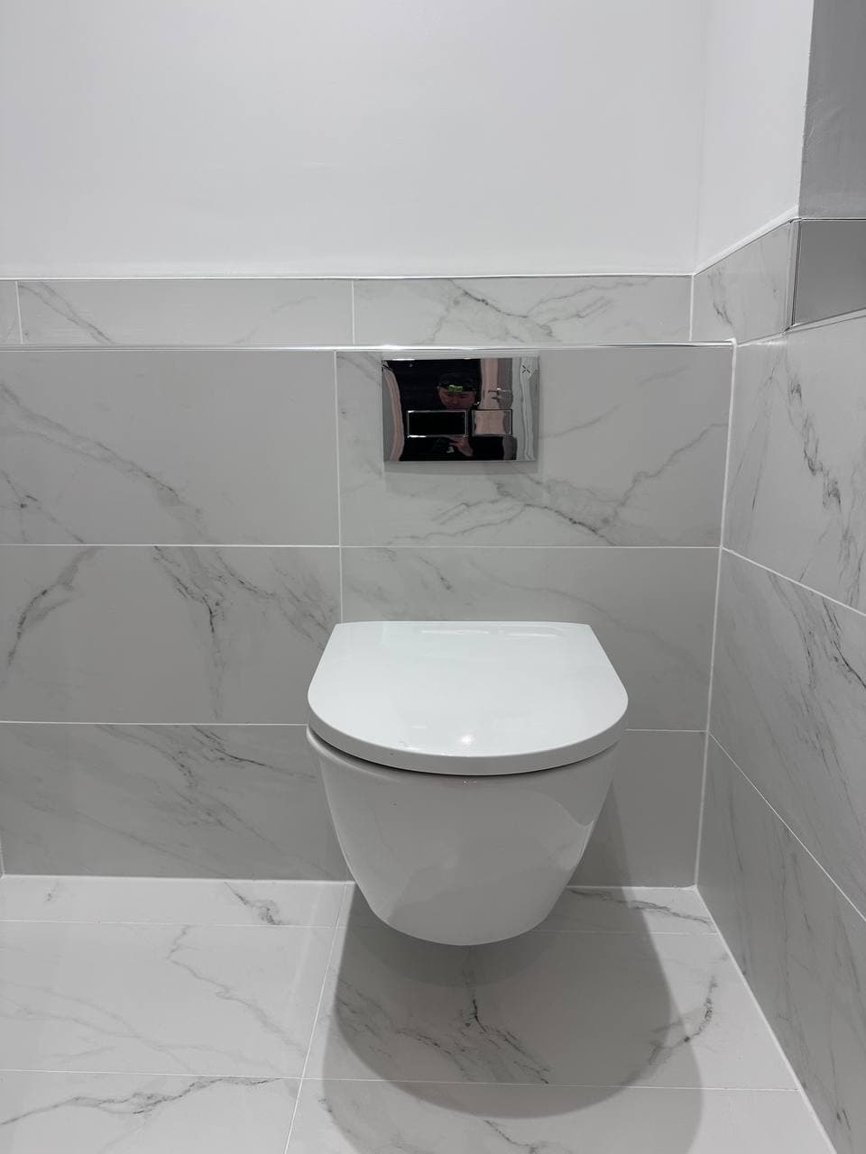 A wall-hung toilet with a chrome flush plate on marble-effect tiles