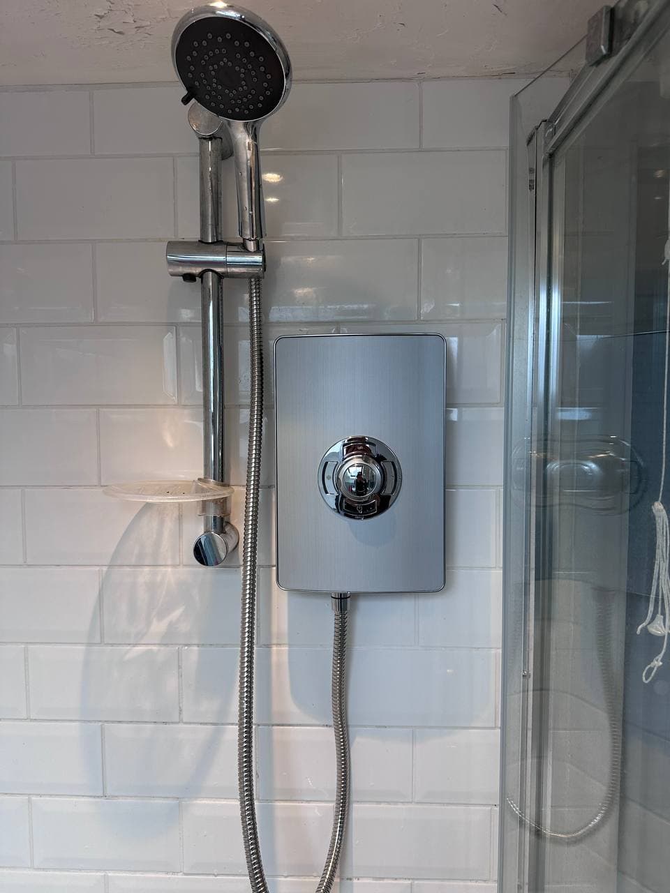 An electric shower and riser rail on white tiles, beside a glass shower door