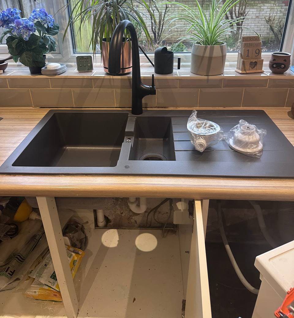 A black kitchen sink and tap fitted in a wooden worktop, with the cupboard doors below still off