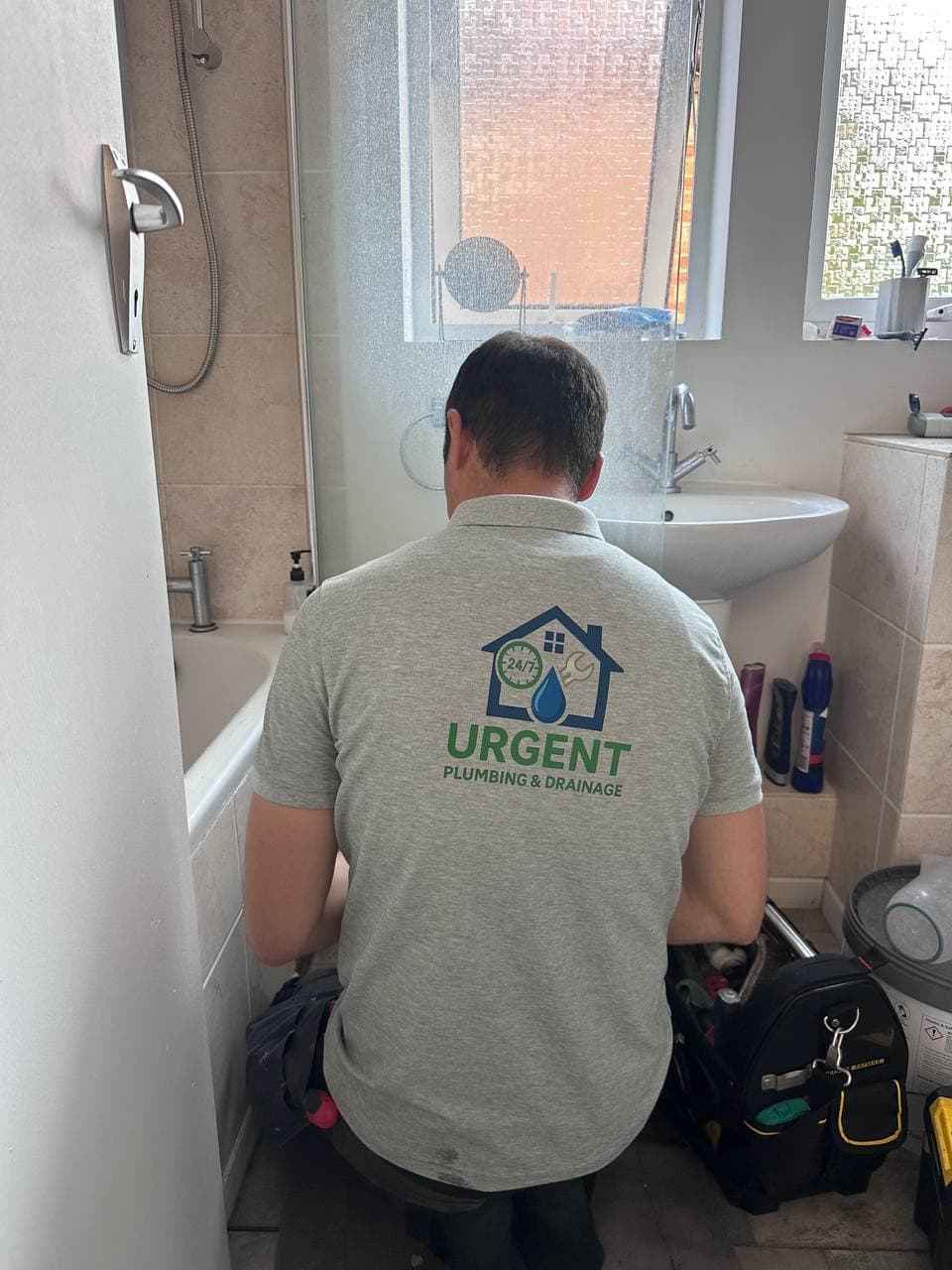 A plumber in an Urgent Plumbing & Drainage polo shirt kneeling beside a bath, with a tool bag, in a customer's bathroom